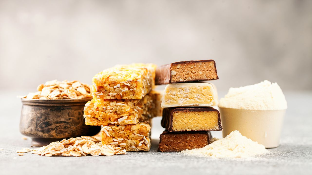 A stack of granola bars next to clean protein bars, with a bowl of oats and a scoop of protein powder on a gray surface.