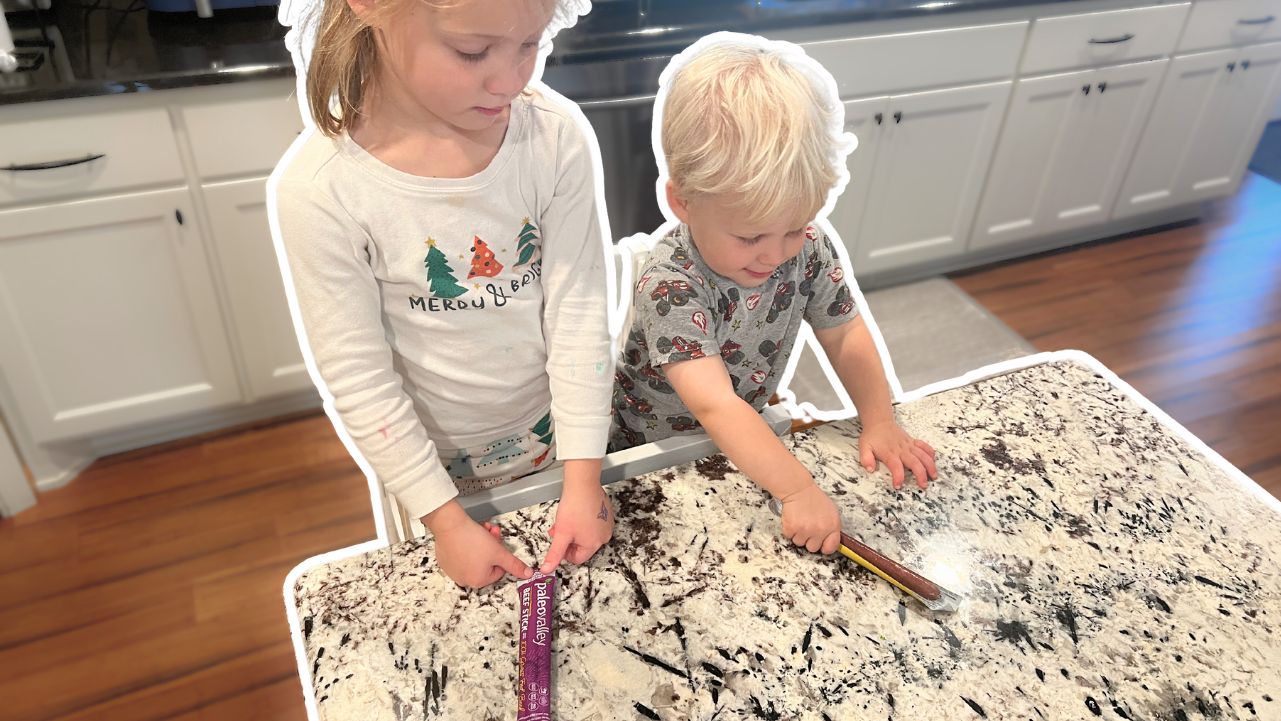 Two young children standing at a counter holding Paleovalley meat sticks.