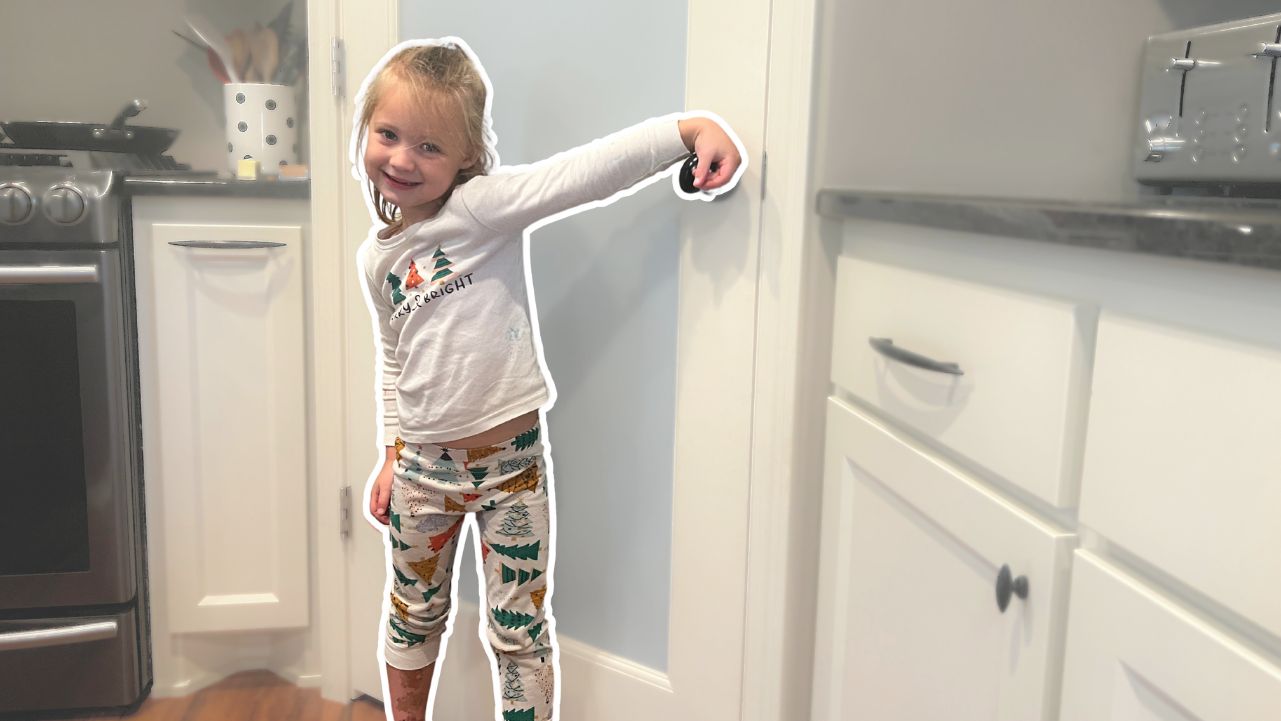 A little girl standing in the kitchen with her hand on a doorknob.