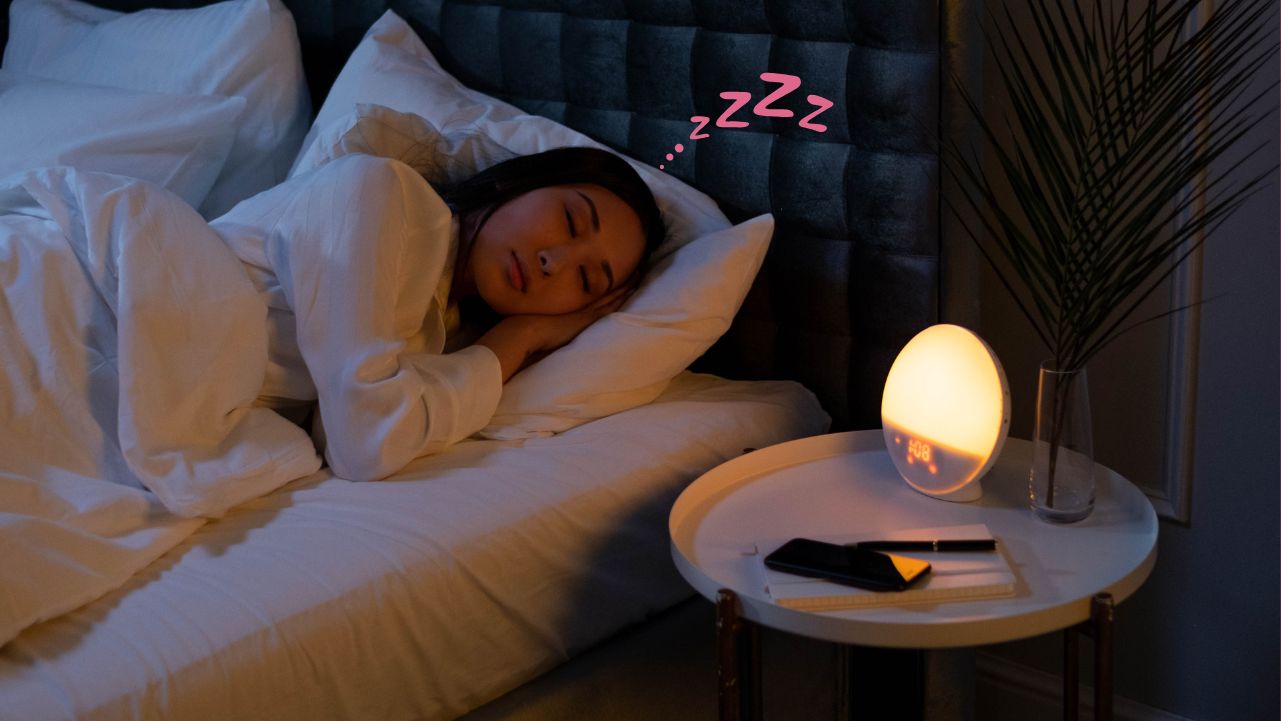 A woman sleeping in a darkened room with a sunrise alarm clock on the bedside table.