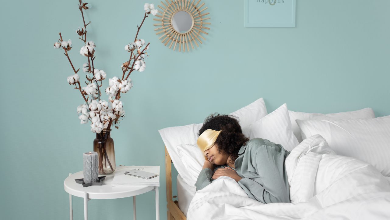 A woman wearing a sleep mask sleeping in a comfortable bed beside a table with flowers and a candle.