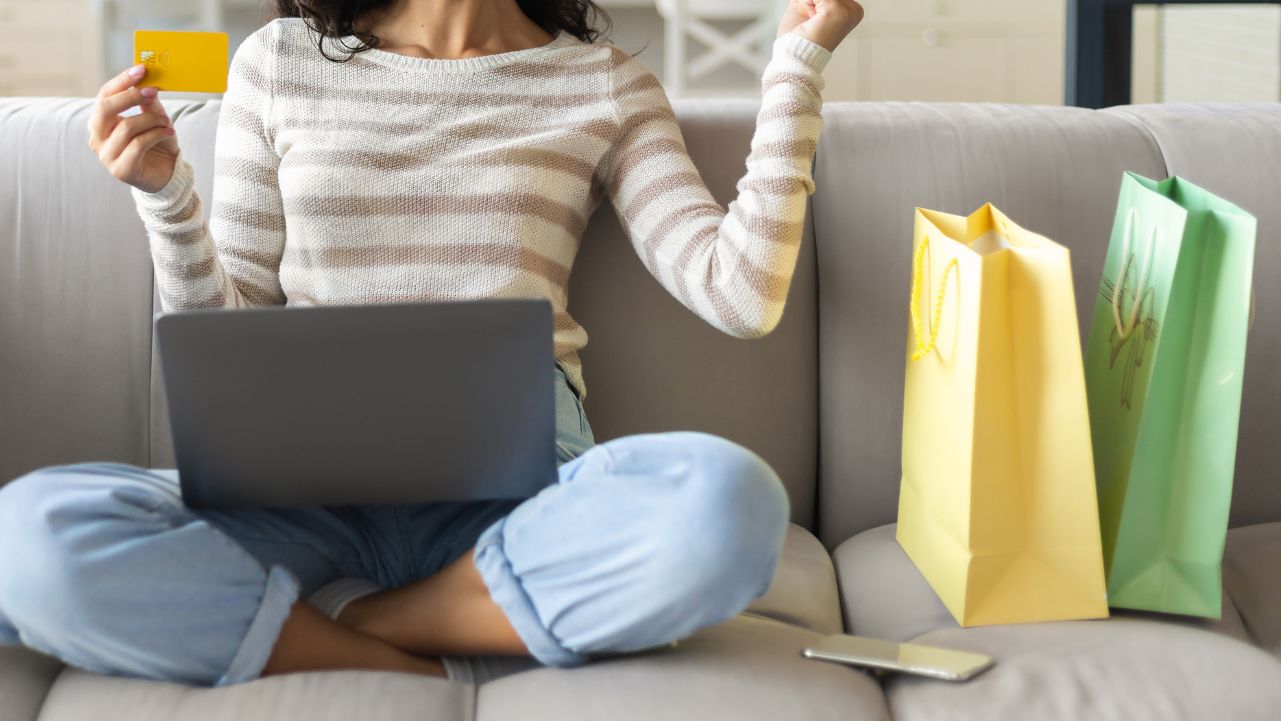 A woman sitting cross legged with a laptop on her lap and a credit card in her hand and two shopping bags on the couch next to her.