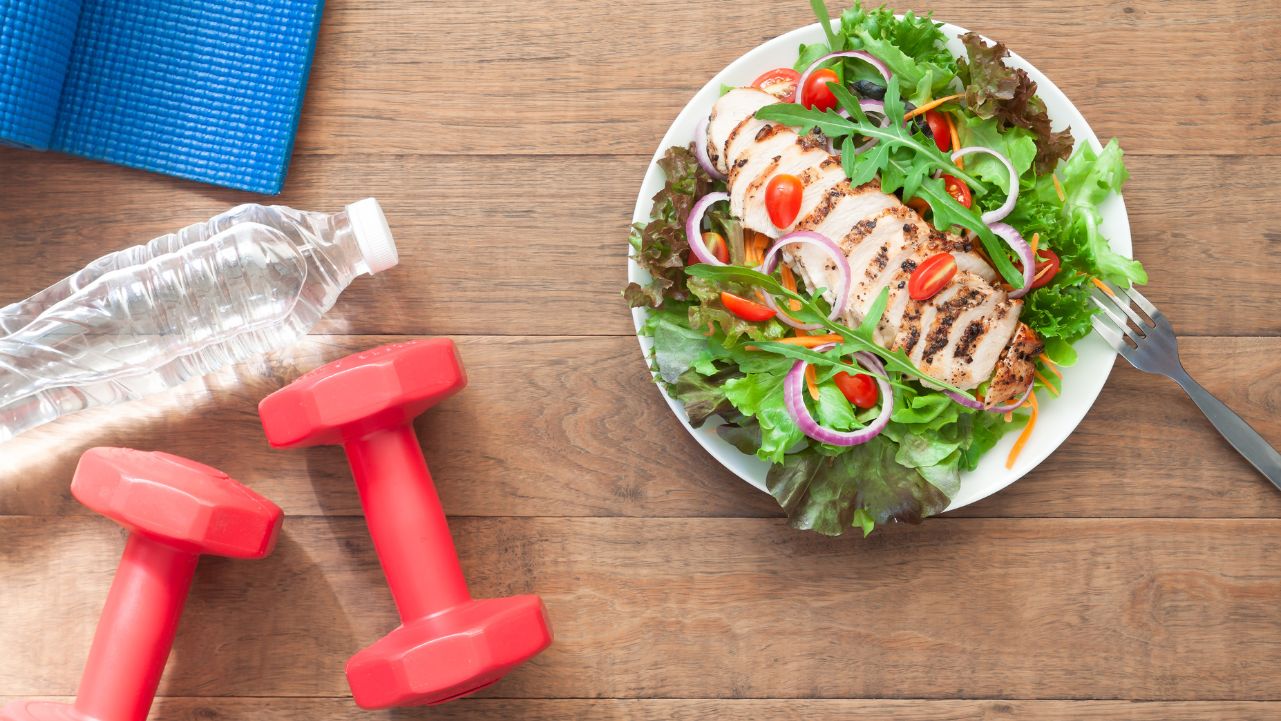 Reset Your Wellness Routine A plate of salad with grilled chicken next to red hand weights, a yoga mat, and a water bottle.