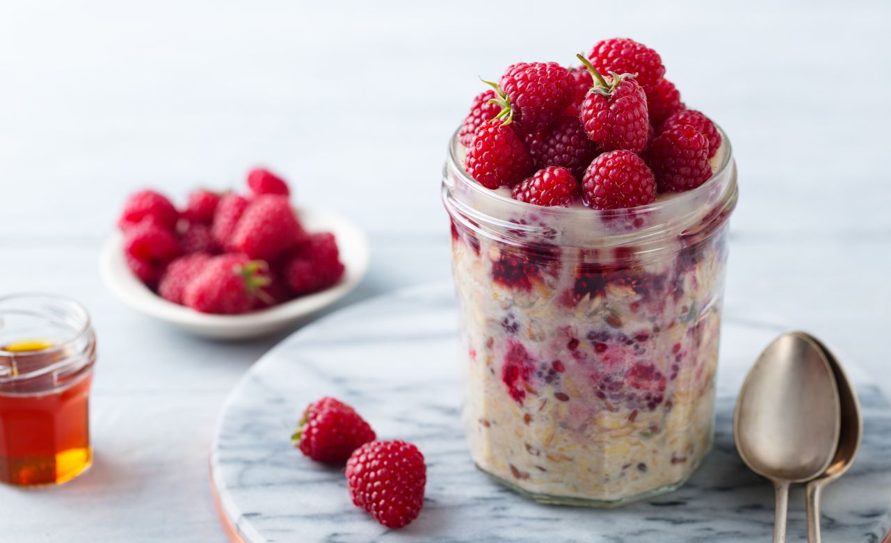 A jar of overnight oats with raspberries, honey, and two spoons