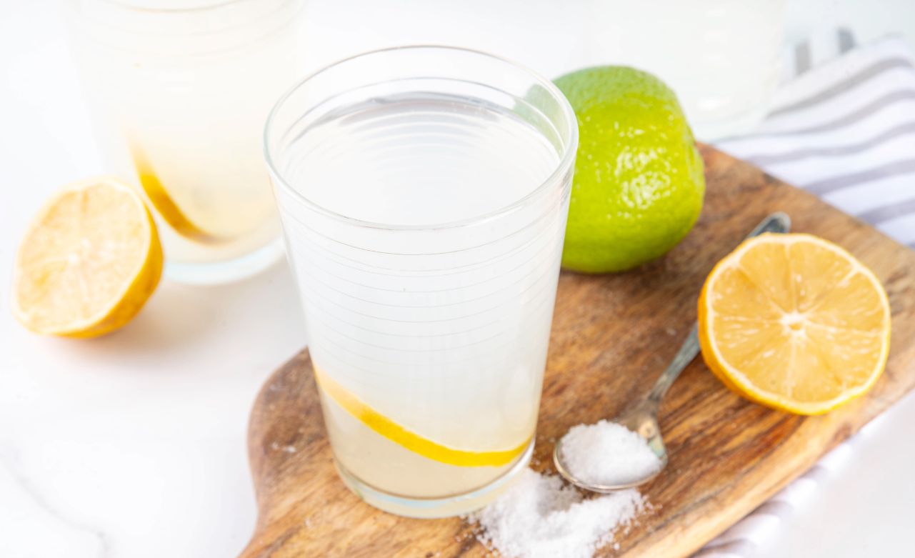 A glass of water, a spoon, a lemon, a lime, and electrolyte salt on a cutting board