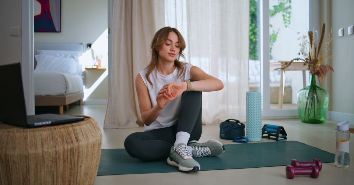 A woman sits on a workout mat at home with exercise equipment and a water bottle nearby after a workout.