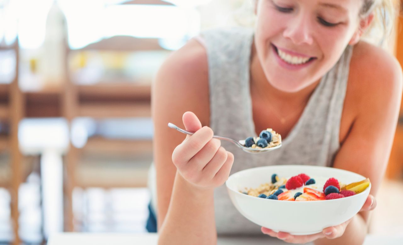 woman-eating-balanced-meal A woman leaning on a counter eating a bowl of fruit and yogurt.