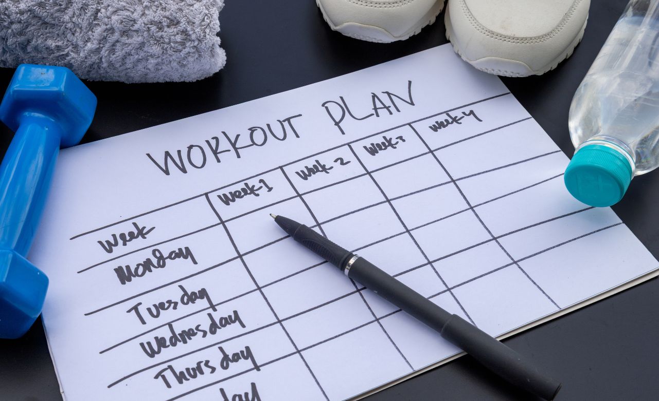 weekly-workout-plan-for-cardiovascular-endurance A paper workout schedule, a pen, sneakers, a blue dumbbell, and a bottle of water.